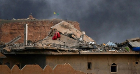 Cette photo prise le 22 mars 2019 montre un toit endommagé et une fumée noire s'élevant du village de Baghouz en Syrie avec des drapeaux de forces kurdes levés.